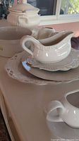 Photo shows white ceramic tureen with lid and handle, two gravy boats with detailed embossed edging, and a set of serving plates with intricate pierced edges and floral designs.
