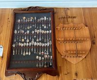 Two wooden display boards on a wooden surface. One board has collector spoons mounted behind glass, arranged in multiple rows. The other shield-shaped board is empty with holders for spoons.