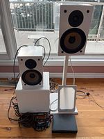 Photo showing two white NHT SuperOne AS1 speakers with black speaker cones, one on a stand and one on a subwoofer, including cables and black audio device on floor.