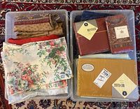 Two plastic bins containing assorted linens including floral printed sheets, tan and red linens, and sealed packages from Revelle and Rumplens brands.