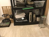 Photo showing black plastic shelving unit with roasting pans and metal canisters on bottom shelf, glass beverage jar with spigot on floor next to shelf.