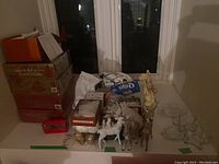 Photo of stacked Christmas decoration boxes, reindeer figurines, and various items on a table near window.