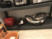 Shelf holding multiple pans including a red Dutch oven, brown frying pan, and black pan under Starfrit Giant Cooker with glass lid