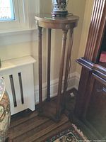 Tall wooden plant stand with round top, four round legs and decorative rosettes, positioned near a window and furniture.