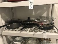 Multiple pots and pans set on a white plastic shelf including black handled pans and glass lids