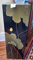 Front side of the black lacquered stand showing gold and green floral designs with large leaves and a lotus bud.
