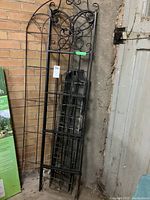 Photo of several black metal garden trellises and gate-style fence sections leaning against a brick wall and concrete floor in a garage. The trellises have scrollwork and arched tops while the fence panels have rectangular grid designs.