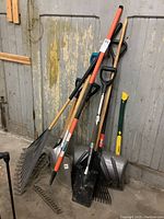 Overall photo showing yard tools leaning against wooden wall including shovels, rakes, and a snow shovel