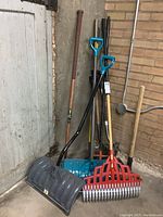 Photo showing all yard tools grouped together, including shovels, rakes, broom, and pickaxe leaning against a corner with concrete floor and brick wall.