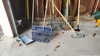 Lot of garden tools standing on a concrete floor including rakes, a blue plastic leaf scooper, a garden hoe, and a pole sander.