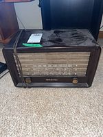 Front view of a vintage RCA Victor tube radio with large dial and two knobs, brown casing dusty on top, showing tape with lot number on top left.