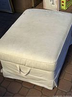 White ottoman with fabric cover, removable top, and side handle for opening storage inside, slight discoloration and wear visible.