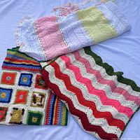 Three folded crochet Afghan-style blankets shown on white background: one with rainbow square motifs, one pastel striped, and one multicolor chevron striped.