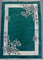 Full view of a large rectangular rug with green central area and cream border, decorated with floral patterns, showing a visible stain in the center area.