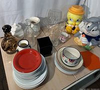 Photo showing Looney Tunes cookie jars, glass decanters, vases, ceramic plates and bowls, mixed glasses, mug and heat pad on a wooden surface under natural light.
