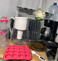 Photo showing various kitchenware items including a drip coffee maker, water filter pitcher, glass plates, silicone baking mold, pie tin, coffee filters, and measuring spoon.