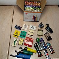 Full lot overview with box of colorful candles behind assorted matchbooks and lighters arranged in front on table