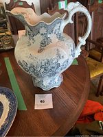 Large antique porcelain water pitcher displaying blue floral patterns and scrollwork with ornate handle and ruffled rim, positioned on wooden table.