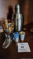 Photo of a cocktail shaker, glass boot-shaped glass with gold floral design, owl-shaped shaker, and a glass shot glass on a wooden surface.