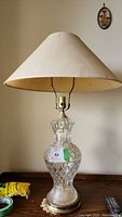 Clear full view of the lamp showing glass body, brass base, and beige fabric shade on wooden surface.
