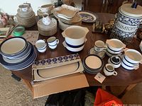 Wide view of all porcelain dinnerware and serveware pieces displayed on wooden table showing plates, cups, pitchers, and a labeled lidded ceramic jar.