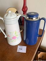 Two insulated pitchers on wooden table: one white ceramic with floral design, the other blue plastic with metal ring near top.