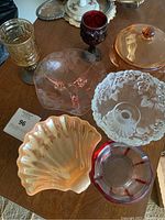 Wide view of assorted vintage glassware including carnival glass shell dish, clear embossed bowl, yellow pressed glass tumbler, ruby red goblet, pink depression glass bowl, and amber covered dish all on wooden table.