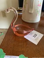 Pink Murano glass swan figurine with a hollow bowl-like body, shown on a table next to a teapot.