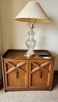 Pressed wood cabinet shown with a crystal lamp and a remote control on top. Geometric X-shaped trim on doors and metal handles visible. Cabinet appears medium brown with some dust.