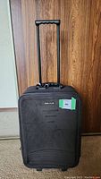 Front view of upright black suitcase with telescoping handle extended, showing brand logo and green tag.