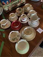 Overview photo showing nine assorted teacups with matching saucers arranged on wooden table, showcasing various patterns including floral and gold trim