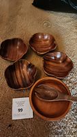 Photo showing five wooden salad bowls and a wooden serving spoon placed on carpet. The bowls include one large round bowl and four smaller curved petal-shaped bowls. Visible wood grain and warm brown color.