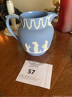 Blue jasperware pitcher with white relief figures and garlands on a wooden table with auction tag 57