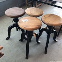 Four adjustable stools arranged on floor, showing round wooden seats and black metal bases with screw height adjusters.