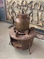 Cast iron cook stove with copper kettle placed on top, showing entire cooking apparatus and kettle handle.
