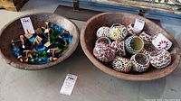 Two large wooden bowls side by side on floor. Left bowl holds blue and green glass and metal hardware items. Right bowl holds various colorful ceramic or mosaic pots.