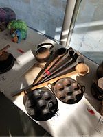 Photo showing the full lot on a white table including 5 wooden spoons, metal teapot, and two cast iron pans with six indentations each