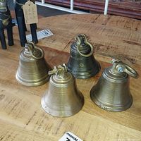 Four vintage brass bells arranged on a wooden table showing the tops and body of each bell, highlighting their brass material and ring handles.