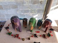 Five hand carved and painted wooden elephants in various colors arranged in a row with the elephant bell garland laid in front of them