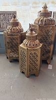Three brass tone Moroccan style punched tin candle lanterns with domed tops and hanging loops, standing on floor against a wall.
