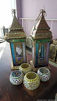 Photo showing 2 Moroccan style punched tin hanging lanterns with colored pressed glass panels in purple, blue and amber tones, surrounded by 4 mosaic glass candle holders in two different patterns.