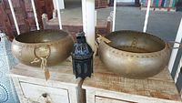 Two large oval metal bowls with rivets and handles on a wooden table, small black tin candle lantern with glass panels between them