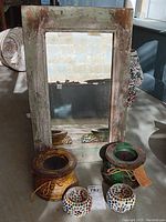 Rustic wood framed mirror with visible distressed paint finish along with two wooden candle holders and two glass mosaic votive holders placed below.