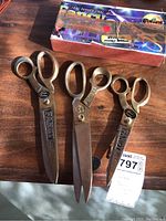 Three pairs of vintage steel scissors laid out on wood surface with original Fancy branded box in background.