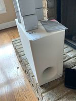White rectangular Bose Acoustimass 7 subwoofer with bass port, sitting on stone hearth near fireplace. Shows size and port detail.