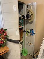 Plastic Rubbermaid cabinet partially open revealing four shelves and pegboard on inside of door holding gardening tools and sun hat.