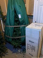 Full view of tree stored inside a green tall storage bag with brown straps, standing upright next to the original box in a basement closet space.