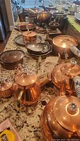 Overview of copper kitchen items including teapots, kettle, coffee grinder, and sauté pans arranged on a granite countertop.