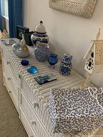 Wide view of multiple blue and white ceramics, glass pieces, decorative birdhouse, and covered box on a wicker cabinet.