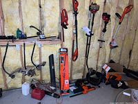 Wide view showing a variety of gas-powered and electric trimmers, hedge trimmers, attachments, and a red gas can arranged against basement wall insulation.